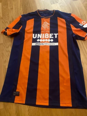 Randig Rangers matchtröja Castore - Snygg Rangers matchtröja från Castore med vertikala marinblå och orange ränder. Tryck med UNIBET och Zero% Mission på framsidan, samt TAVERNIER och nummer 2 på ryggen. Korta ärmar och sportig passform, perfekt för fotboll eller träning.