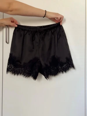Svarta satinskjorts med spetskant - Snygga svarta loungewear-shorts i satin med elastisk midja, snörning. Shortsen har dekorativ spetskant längst ner som ger en lyxig och feminin känsla. Helt nya utan prislapp, endast testade (lite för små). Nypris 369kr. 