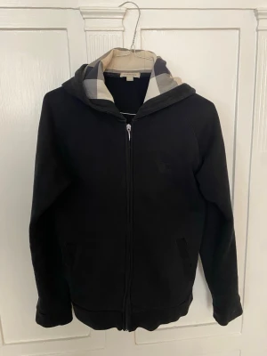 Svart hoodie från Burberry med dragkedja - Svart hoodie från Burberry med klassiskt rutigt foder i huvan och dragkedja framtill. Storlek 14y/162cm