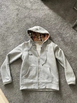 Grå hoodie från Burberry med dragkedja - Snygg grå hoodie från Burberry med klassiskt rutigt foder i huvan och dragkedja framtill. Har två stora fickor och broderad logga på bröstet. Perfekt för en chill och stilren look. Tillverkad i mjukt bomullsmaterial.