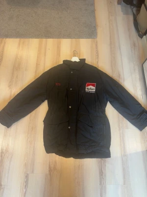 Svart pufferjacka från Hugo Boss - Säljer en svart pufferjacka från Hugo Boss med Marlboro och Team Penske Sponsor patch på bröstet. Jackan har hög krage, tryckknappar, två stora fickor framtill och broderad BOSS-logga. Perfekt för kalla vinterdagar och riktigt snygg streetstil.
