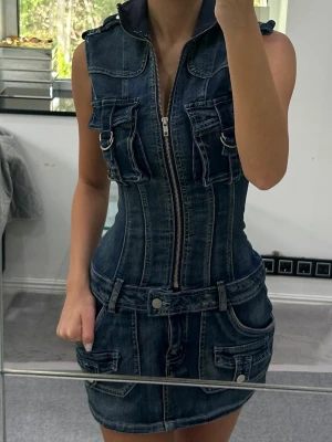 Ärmlös jeansklänning med dragkedja - Säljer en cool ärmlös jeansklänning i mörkblå denim med markerad midja och flera fickor fram. Klänningen har dragkedja hela vägen framtill. Den har fickor bak