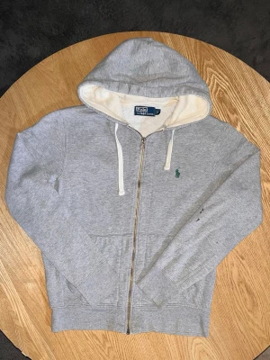 Polo Ralph Lauren Zip Hoodie Grå – Storlek S - Säljer en Polo Ralph Lauren zip-hoodie i grå melange.  Klassisk modell med broderad Polo-logo på bröstet, huva med vita snören och praktiska fickor framtill.  Storlek: S Skick: Bra. Tvättad och klar. Lite fläckar/märken syns på vänster arm (se bilder), annars fin.  Unisex: Ja Mått: Kan tas vid förfrågan.