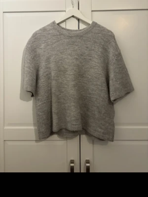 Grå stickad kortärmad tröja - Mysig grå stickad tröja med korta ärmar och rund hals. Tröjan har en loose passform och ribbade kanter vid ärmslut och nederkant. Perfekt att styla med jeans eller kjol för en avslappnad look.