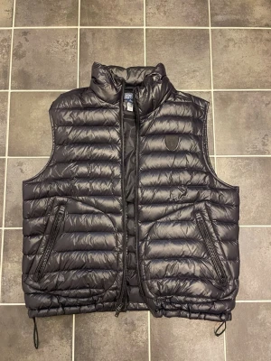 Polo Ralph Lauren glossy väst  - Svart Polo dunväst med hög krage och två dragkedjeförsedda fickor framtill. Västen har quiltad design med horisontella sömmar och justerbar dragsko nedtill. Perfekt för lager-på-lager och enkel att matcha till olika outfits. Snygg och perfekt till hösten! 