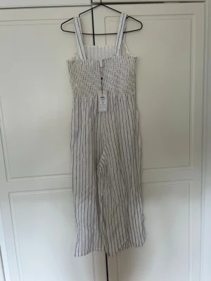 Vit jumpsuit med blå ränder - Säljer en snygg vit jumpsuit med tunna blå vertikala ränder. Jumpsuiten har breda axelband, smockad överdel och raka, vida ben. Tillverkad i ett lätt tyg som känns luftigt och perfekt för sommaren. Enkel och stilren design som är lätt att matcha.