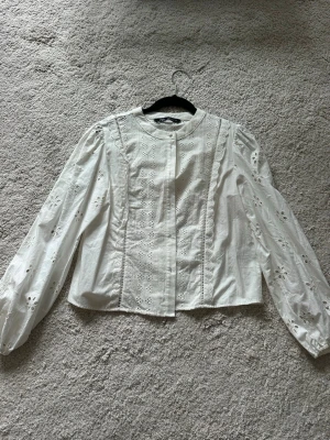 Vit blus med broderade detaljer - Söt vit blus från Zara med långärmade ärmar och broderade hålmönster. Blusen har rund halsringning och knäppning framtill. Tillverkad i ett lätt bomullsliknande material som känns luftigt och fräscht. 