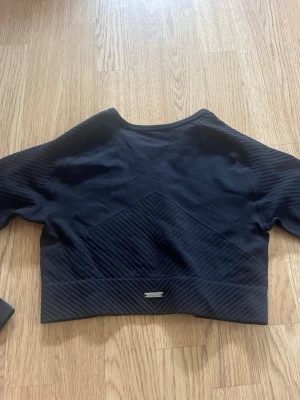 Svart sporttröja med strukturmönster - Säljer en svart sporttröja med snyggt strukturmönster och tumhål i ärmarna. Tröjan har en croppad passform och är tillverkad i ett stretchigt material som passar perfekt till träning eller gym. Metallogo framtill.