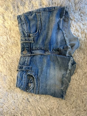 Blå jeansshorts med rå kant - Säljer ett par blå jeansshorts med rå, fransig kant och coola detaljer. Shortsen har klassiska fickor fram och bak, bälteshällor och knappstängning. Perfekta för varma dagar och ger en avslappnad vibe.