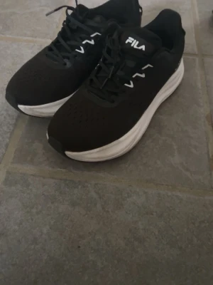 Svarta FILA sneakers med vit sula - Säljer ett par svarta sneakers från FILA med vit, tjock sula och svarta skosnören. Skorna har ventilerande meshmaterial upptill och vita detaljer vid snörningen. Perfekta för dig som gillar sportig och clean stil.