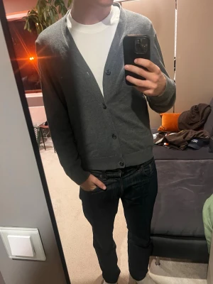 Mörkgrå cardigan från Pier one - Stilren mörkgrå cardigan från Peak One med v-ringning och knäppning framtill. Tröjan har långa ärmar och ribbade muddar vid ärmslut och nederkant. Perfekt att slänga över en t-shirt för en clean look.