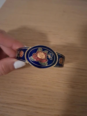 Blått armband med blommotiv - Snyggt armband i metall med djupblå emalj och detaljerade blommor i rött, rosa och guld. Ovalt mittparti med rosdesign och guldfärgade kanter. Armbandet har gångjärn för enkel öppning och stängning. Perfekt för dig som gillar färgstarka accessoarer.