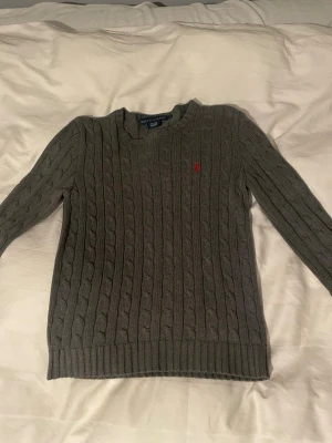 Grå kabelstickad tröja Ralph Lauren - Snygg grå kabelstickad tröja från Ralph Lauren med rund halsringning och lång ärm. Tröjan har det klassiska röda Polo-logotypen broderad på bröstet och ribbade muddar vid ärmslut och nederkant. Perfekt för lager på lager och stilren look. Sitter typ som en xs