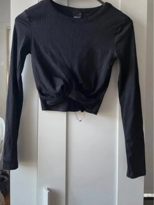 Svart ribbad crop top med twist - Snygg svart långärmad crop top i ribbat material med twistad detalj framtill. Tröjan har rund halsringning och figurnära passform, perfekt till höga jeans eller kjol. Enkel men trendig design som passar till många olika stilar.