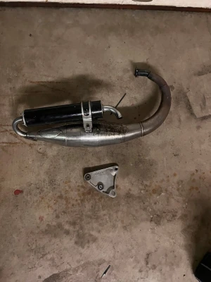 Avgassystem till moped - Säljer ett avgassystem till moped med svart ljuddämpare och böjt rör i metall. Medföljer även en monteringsplatta. Passar till dig som vill byta eller uppgradera ditt mopedsystem. Perfekt för dig som gillar att meka.