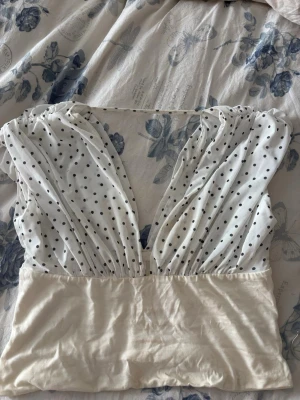 Vit prickig omlott topp - Snygg vit omlott topp med djup v-ringning och prickigt mönster på överdelen. Toppen är ärmlös och har en croppad passform som ger en trendig look. Perfekt att matcha med höga jeans eller kjol för en cool stil.