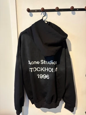 Svart hoodie Acne Studios Stockholm - Svart oversized hoodie från Acne Studios med tryck i vitt på ryggen där det står 'Acne Studios STOCKHOLM 1996'. Klassisk huva och ribbade muddar vid ärmslut och nederkant. Perfekt för en avslappnad streetwear-look.