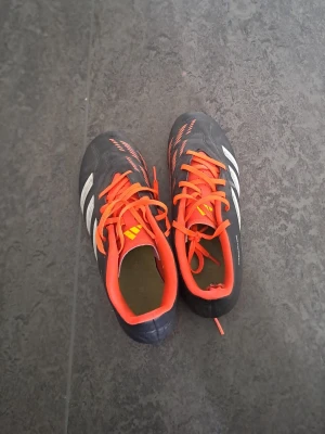 Adidas Predator fotbollsskor svart/orange - Säljer ett par Adidas Predator fotbollsskor i svart med vita detaljer och orange snörning. Skorna har klassisk design med tre vita ränder på sidan och orange insida. Perfekta för dig som vill sticka ut på planen!