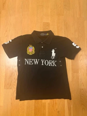 Svart pikétröja New York Polo - Svart pikétröja från Polo Ralph Lauren med vit broderad logga, texten NEW YORK framtill och siffran 5 på ryggen och ena ärmen. Tröjan har krage, knappar och detaljer i gult och vitt på bröstet.