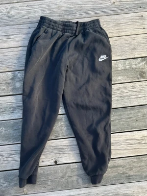 Svarta mjukisbyxor från Nike - Svarta mjukisbyxor från Nike med resår och dragsko i midjan, samt muddar vid bensluten. Byxorna har två sidofickor och en vit Nike-logga på vänster ben. Tillverkade i mjukt och bekvämt material, perfekta för chill eller träning.