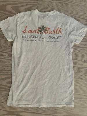 Saint Barth t-shirt  - Säljer en vit t-shirt med färgglatt tryck på ryggen där det står 'Saint Barth Billionaire's Resort' och en palm i mitten. T-shirten har rund hals och korta ärmar. Perfekt för dig som gillar avslappnad och trendig stil.
