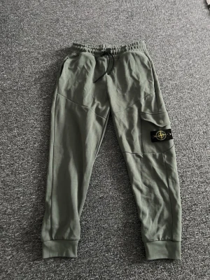 Gröna mjukisbyxor med logga - Snygga gröna mjukisbyxor med resår i midjan och dragsko. Byxorna har en ficka på benet med en svart patch och gul logga, samt ribbade muddar vid bensluten. Perfekta för en avslappnad stil.