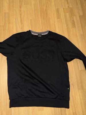 Svart stickad tröja från Hugo Boss - Snygg svart stickad tröja från Hugo Boss med diskret broderad logga på bröstet. Tröjan har rund halsringning och ribbade muddar vid ärmslut och nederkant. Perfekt för en stilren look.