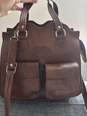 Brun handväska med dubbla fickor - Snygg brun handväska i konstläder med två stora fickor framtill och vågig kant upptill. Väskan har både handtag och avtagbar axelrem med silverfärgade detaljer. Perfekt för dig som vill ha plats för allt viktigt och gillar en klassisk men cool look. Mått: 28x26x10 cm 
