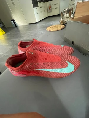 Röda Nike fotbollsskor utan snörning - Säljer ett par röda Nike fotbollsskor med turkos swoosh och modern design utan snörning. Skorna har en strömlinjeformad form och ovandel i syntetmaterial som ger bra bollkontroll och passform. Perfekta för dig som vill sticka ut på planen.