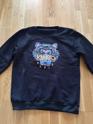 Svart sweatshirt med tigertryck Kenzo - Cool svart sweatshirt från Kenzo med ikoniskt tigerhuvud broderat i färgglada toner på bröstet och texten 'KENZO PARIS'. Tröjan har rund halsringning och ribbade muddar vid ärmslut och nederkant. Perfekt för dig som gillar streetwear och statement-plagg. Står xxl men passar M/L