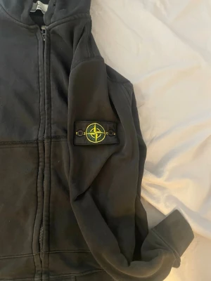 Svart hoodie med dragkedja Stone Island - Svart hoodie från Stone Island med dragkedja och huva. Klassisk patch med logotyp på vänster ärm. Stora fickor framtill och ribbade muddar. Perfekt för dig som gillar streetwear och vill ha något stilrent och bekvämt.