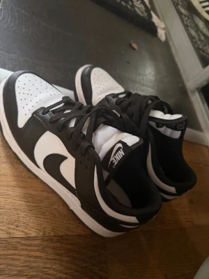 Nike svartvita låga sneakers - Säljer ett par klassiska Nike sneakers i svart och vitt. Skorna har lågt skaft, svart swoosh och svarta detaljer mot vit bas. Ovandelen är i läder med perforerad tåbox för extra ventilation. Perfekta för dig som gillar stilrena och tidlösa sneakers.