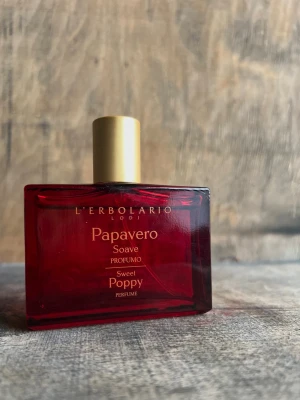 L'Erbolario Papavero ren parfym, 50ml - En elegant parfymflaska i mörkrött glas med guldfärgad spray och lock. Doften heter Papavero Soave Sweet Poppy och kommer från L'Erbolario. Flaskan har en stilren, rektangulär form och text i guld på framsidan.