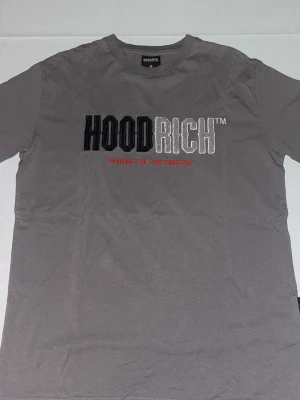 Grå HOODRICH t-shirt med brodyr - Snygg grå t-shirt från HOODRICH i bomull med broderad logga i svart, vitt och rött framtill. Klassisk passform och rund halsringning, perfekt för en streetwear-look.