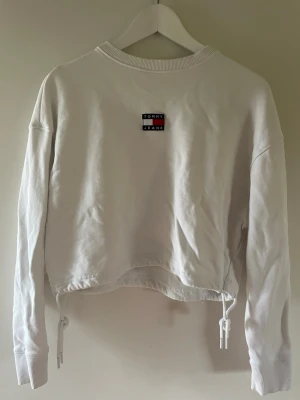 Vit croppad sweatshirt Tommy Jeans - Säljer en vit croppad sweatshirt från Tommy Jeans med broderad logga på bröstet och snörning i nederkanten. Jätte fin och bra skick