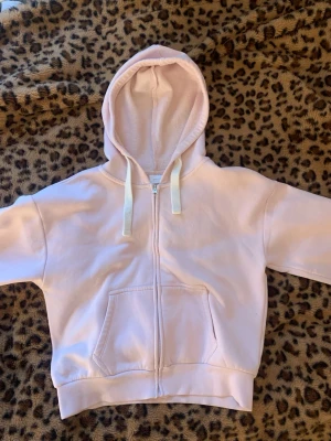 Ljusrosa hoodie med dragkedja - Säljer en ljusrosa hoodie med dragkedja framtill och stora fickor. Hoodien har vita snören i huvan och är gjord i mjukt, fleeceliknande material på insidan. Perfekt för en chill och avslappnad stil.