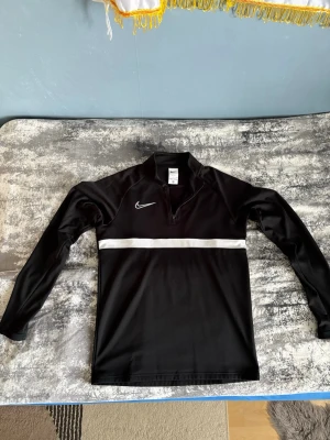 Svart Nike Dri-FIT träningströja - Snygg svart långärmad träningströja från Nike med vit rand över bröstet och halv dragkedja framtill. Tillverkad i Dri-FIT-material som andas och håller dig torr under träningen. Perfekt för löpning, gym eller fotboll.