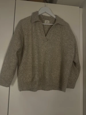 Beige stickad tröja med krage - Mysig beige stickad tröja med bred krage och v-ringning. Tröjan har långa ärmar och ribbstickade muddar vid ärmslut och nederkant. Perfekt för lager på lager och en avslappnad stil.