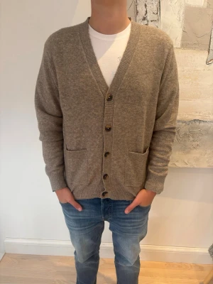 Beige kofta med knappar och fickor - Mysig beige kofta med v-ringning och knäppning framtill. Koftan har två framfickor och ribbade muddar vid ärmslut och nederkant. Perfekt att slänga på sig över en t-shirt för en avslappnad look.
