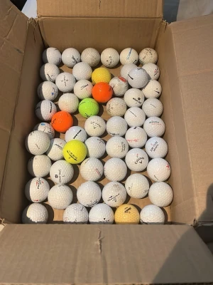Låda med blandade golfbollar - En stor låda fylld med blandade golfbollar i olika färger, mestadels vita men även några gula och orange. Perfekt för dig som vill fylla på golfbagen med många bollar till träning eller spel. Flera olika märken och modeller.