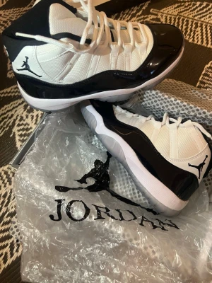 Air Jordan 11 Concord sneakers - Säljer ett par ikoniska Air Jordan 11 Concord sneakers i svart och vitt. Skorna har blankt lackat läder runt om, mesh-tyg på ovandelen och klassisk Jumpman-logga på sidan. Tjock vit sula med isblå detaljer. Perfekta för dig som gillar streetwear och snygg design.