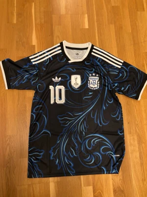 Argentina Messi fotbollströja svart/blå - Snygg Argentina fotbollströja med Messi och nummer 10 på ryggen. Tröjan är svart med blått mönster, vita detaljer och tre vita ränder på axlarna. Framtill finns AFA-logga, FIFA-märke och Adidas-logga. Perfekt för dig som älskar fotboll och Messi.