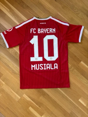 FC Bayern München matchtröja röd - Snygg röd FC Bayern München matchtröja från Adidas med Musiala och nummer 10 på ryggen. Tröjan har vita detaljer, klubbmärke och sponsorlogga framtill samt guldiga inslag vid kragen. Tillverkad i lätt och ventilerande material, perfekt för fotboll eller träning.