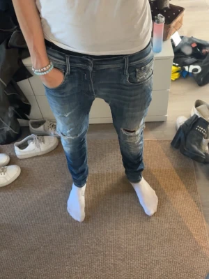 Replay Anbass Jeans  - Säljer ett par blå jeans med låg gren och slitningar på benen. Jeansen har en avslappnad passform, klassiska fem fickor och detaljerade sömmar. Perfekta för en casual och trendig look. Storlek 30/32