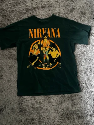 Nirvana t-shirt från Pull&Bear - Säljer en mörkgrön Nirvana t-shirt från Pull&Bear med stort tryck av bandet och loggan i gult framtill. Klassisk rund hals och korta ärmar. Perfekt för dig som gillar bandtröjor och vill ha en cool och avslappnad stil.