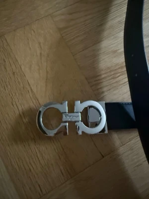 Svart ferragamo bälte med silverfärgat spänne - Stilrent svart bälte med ett iögonfallande silverfärgat metallspänne. Bältet har en klassisk, slät design och passar perfekt till jeans eller kostymbyxor. Spännet har en unik form som ger en exklusiv känsla.