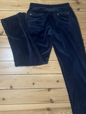 Mörkblå velour mjukisbyxor - Säljer ett par mörkblå mjukisbyxor i mjuk velour med resårmidja och dragsko. Byxorna har stora fickor både fram och bak, samt detaljer med metallknappar på bakfickorna. Perfekta för chill dagar eller när du vill vara extra bekväm.