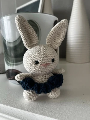 Virkad kanin med blå kjol - Söt handvirkad kanin i beige bomullsgarn med stora öron och svarta ögon. Kaninen har en mörkblå virkad kjol och ett litet broderat rosa leende. Perfekt som dekoration eller present.