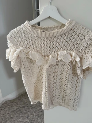 Vit virkad topp med volang - Supersöt beige/vit virkad topp med korta puffärmar och bred volangdetalj över bröstet. Tröjan har ett fint spetsmönster och ribbad halsringning. Perfekt för dig som gillar romantisk och bohemisk stil. Materialet känns mjukt och luftigt, troligen bomull.
