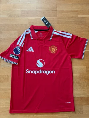 Manchester United matchtröja röd - Säljer en röd Manchester United matchtröja från Adidas med vita detaljer och klubbmärke på bröstet. Tröjan har trycket 'B. Fernandes 8' på ryggen, Premier League-märke på ärmen och Snapdragon som sponsor. Tillverkad i lätt funktionsmaterial, perfekt för fotboll.